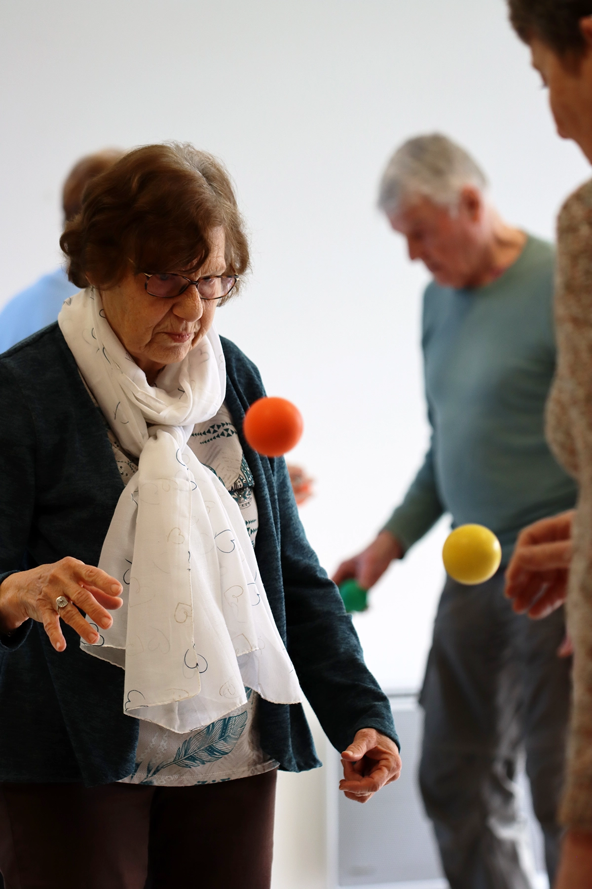 Formation pour kinésithérapeutes : atelier Brain Ball avec seniors ; une femme âgée fait rebondir une balle orange pour travailler rythme, coordination et prévention des chutes (groupe en arrière-plan).